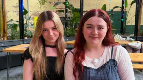 A landscape image of two sisters smiling at the camera - one with long blonde hair and the other with long, lightly curled red hair. The sister with blonde hair is wearing a black top and silver necklace, while the other is wearing a white t-shirt with denim over the top of it. They are sitting at a wooden table on a wooden bench. In the background, more wooden tables and benches can be seen amongst plants and blue and green fairy lights.