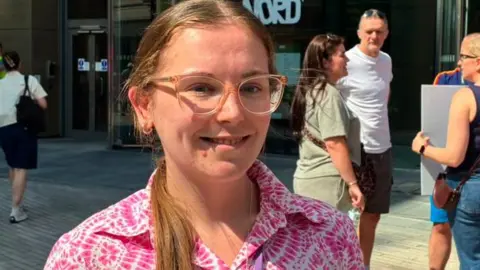 Heather Taylor with long blond hair in a ponytail wearing glasses and a pink and white shirt. She stands outside the Plaza building in Liverpool city centre. She is smiling. Protesters stand behind her.