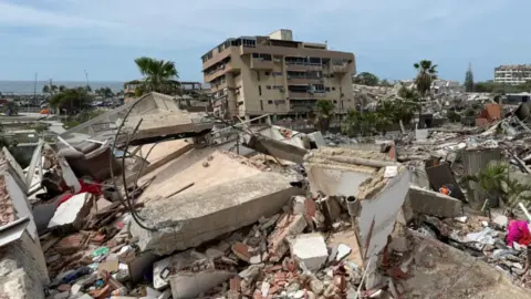 There are huge broken concrete blocks with wire sticking out of them lying on piles of rubble, with smaller pieces of debris visible underneath. A block of flats in the background is still standing but appears to be leaning slightly. to the right of the picture a rescue worker wearing an orange high visibility top appears dwarfed by the broken buildings around them.