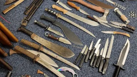 IOM Constabulary A range of sharp and serrated knives laid out on a blue carpeted floor. 