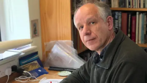 Tim Noad sits at his desk - he is looking at the camera from the right, and wears a dark fleece. There's a bookcase in the background.