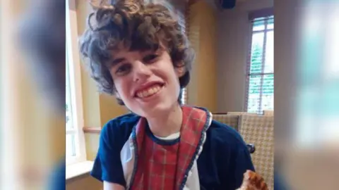 Family handout A boy with brown curly hair giving a teethy smile at the camera and wearing a black top and red bib. He is holding a piece of toast in his hand.