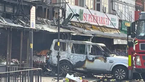 A burnt-out vehicle, with charred bodywork, stands near the scene of the fire and a fire engine can be seen to one side.