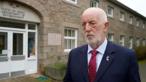 Steve Luce in a navy blue suit and red tie. He is standing outside the environment department. 