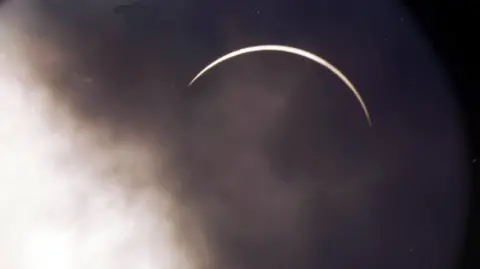 PA Media The moon is covering the sun, leaving a fingernail of light. The cloud over the moon is dark and there is lighter cloud to the left.