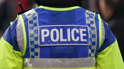 A police officer with his back to the camera wears a yellow and blue high-vis jacket with the word 'police' on the back. 