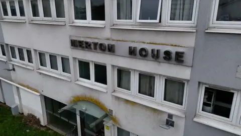 A close-up view of a white tower block showing metal lettering reading 'Merton House' mounted on the facade. The building shows signs of weathering with yellow-green staining around the entrance canopy and lettering. Several rows of white-framed windows are visible above and below the sign.