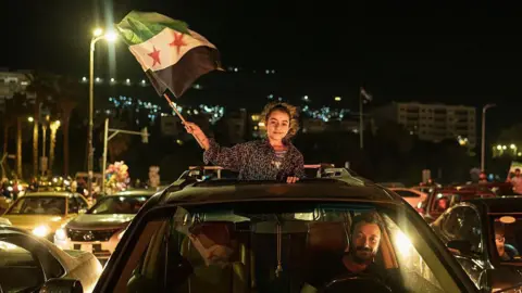 A girl waves a Syrian flag out of the open roof of a car being driven by a smiling man in Damascus