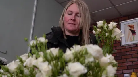 Woman with blonde hair in a black coat and a black top is picking white roses out of a large flower collection.