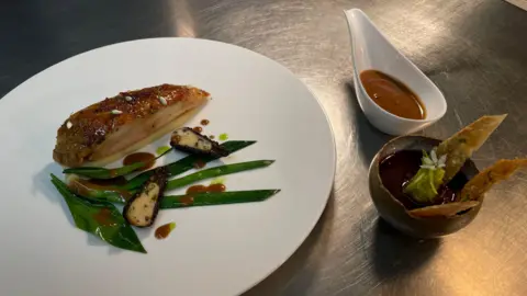 Plate with crispy chicken, leeks, wild garlic and morels decorated with a jus and gravy like sauce. Next to the plate, there are two more dishes, one small jug with the jus in and a small bowl with crispy chicken skin and chicken liver pâté.