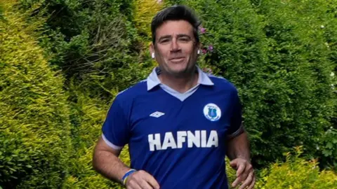PA Media Andy Burnham, who has dark hair, photographed while running. He is wearing a retro blue Everton FC shirt from the 1980s.