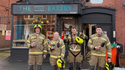 Tiny Bakery Four firefighters in full protective gear, stood outside the shop holding coffees and pastries
