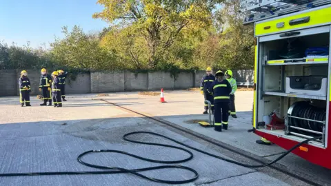 A fire truck is seen to the right with a partly-uncoiled hose, while there are two clusters of firefighters, one in the middle ground and one in the background, all dressed in full firefighter gear. 