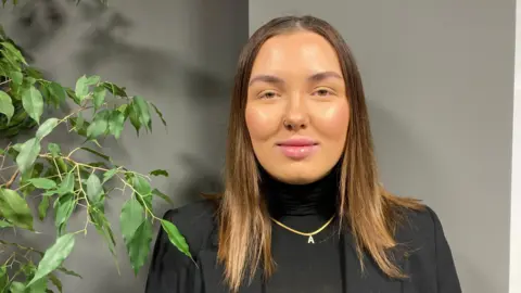 Chelsea Hutton, wearing a black top and blazer. She is wearing a gold necklace with the letter A. She has short brown hair. She is stood in front of a grey wall. 