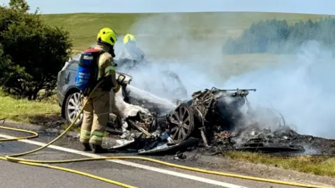 Smoke emerges from a car on fire as firefighters tackle the fire.