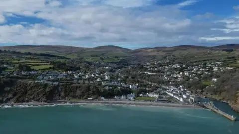 An aerial view of Laxey, which shows buildings on hilly terrain with a long arching coastline in front of turquoise waters.