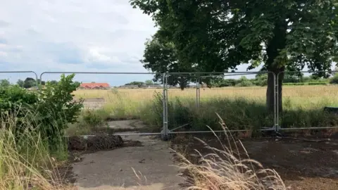 Temporary metal fencing is in place in front of an overgrown field, with long grasses and plants. There are patches of concrete on the ground, which are overgrown with moss and dead plants.