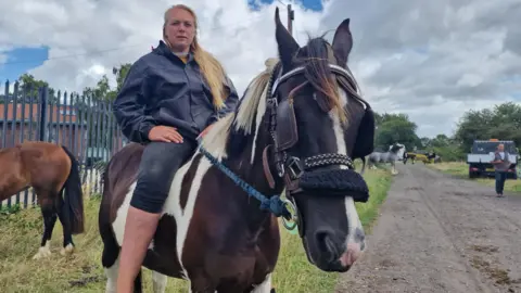 BBC Woman with long blonde hair, blue jacket and jeans, sitting on a horse. There is another horse in the background, some grass and a track with more horses, a vehicle and a person.