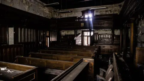 Pugh Auctions A dilapidated courtroom. There are a number of dusty wooden benches in the partially wood-panelled room. Peeling paintwork can be seen above the dark panelling.