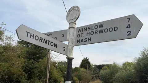 Amy Holmes/BBC Traditional signpost at a rural crossroads in Beachampton, Buckinghamshire, pointing to Thornton, Winslow, Great Horwood, Nash and Thornborough along a country lane.