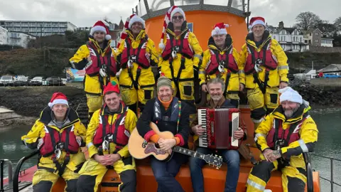 Eight RNLI lifeboat crew surrounding Devon songwriters and musicians James Studholme and Chris Hoban, from Police Dog Hogan, as they play their Christmas single 'Pull Away' - in front of an RNLI lifeboat. One of the musicians is holding an acoustic guitar, while the other is playing an accordion. The lifeboat crew are in full red and yellow uniform, with lifejackets on.