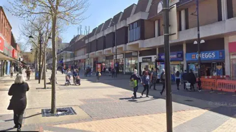 A pedestrianised shopping street. It is a bright and sunny day and there are shops on either side of the street and people walking around. 
