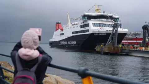 PA Media The Glen Sannox ferry arriving in Brodick on Arran from Troon on its first official day in service with the catamaran Alfred in the background. A person wearing a pink beanie hat, a grey and pink rucksack and a black jacket takes a photograph of the ferry in the foreground of the shot.