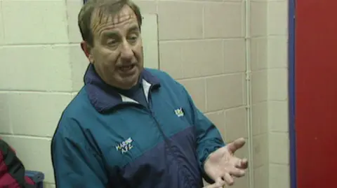 BBC Roly Howard, vestido de turquesa, está en el vestuario de los futbolistas con las manos extendidas. Tiene el pelo castaño corto. 