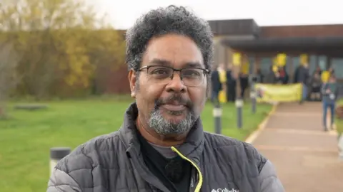 A man in a black jacket with grey sleeves with a yellow zip. He is stood outside, in the forefront of a protest, smiling at the camera