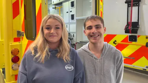 Students Millie and Alfie are standing in front of a teaching ambulance. They are both wearing sweaters and are smiling at the camera.