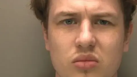 A police custody shot showing a close-up of the face of a man looking directly at the camera. He has brown hair and blue eyes and wears a serious expression. 