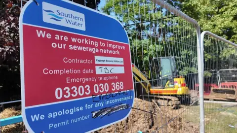Metal fencing with diggers behind and large Southern water sign attached to it.