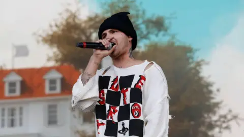 Slowthai performing with a microphone in his hand. He is wearing a black beanie hat and a ripped white shirt.