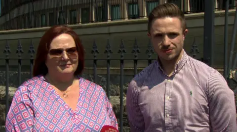Kathryn McCollum has reddish-brown hair down to her shoulders and is wearing a patterned blouse. Adam McCollum has short dark hair swept back from his forehead and is wearing a lightly striped shirt