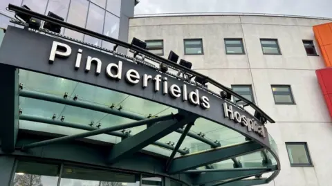 The image shows the exterior entrance of Pinderfields Hospital. A large glass canopy extends over the doorway, supported by metal framework. Mounted above the canopy is the hospital’s name in bold lettering. Behind it, a curved section of the building with multiple windows is visible.
