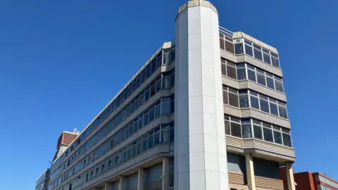 Owen Sennitt/BBC Sovereign House, taken from the southern side up close, looking upwards, showing a cylindrical tower connected to the side of the concrete office block