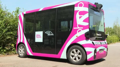 A driverless miniature bus with pink and white livery parked in a car park. 