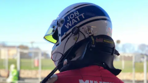 Jon Watt A close up of the back of Jon Watt's head. He is wearing a white helmet with Jon Watt written on it and is at the Brands Hatch circuit.