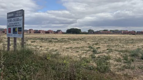 A large piece of grassy land with new build homes in the far distance. A sign advertising the employment space sits on the left side