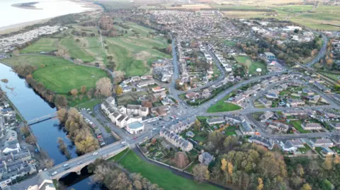 An aerial view of the east coast town of Nairn. There are clusters of houses and green fields. A bridge carries traffic over a river. 