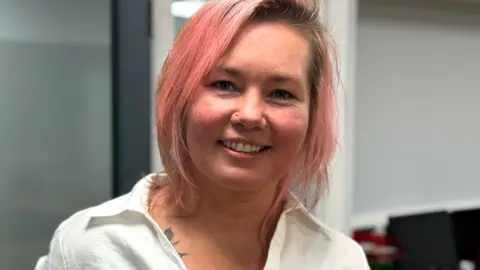 A woman with pink hair is smiling into the camera. She is wearing an open collar white shirt. She has a nose ring and part of her tattoo appears under her shirt.