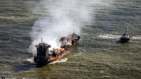 PA Media Tug boats shadow the Solong container ship as it drifts in the Humber Estuary, off the coast of East Yorkshire following a collision with the MV Stena Immaculate oil tanker. Smoke can be seen billowing from the ship which has char and burn marks on it. 