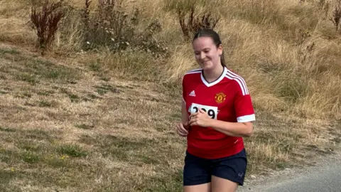 A woman running and wearing a red adidas t shirt and black shorts and smiling s she runs. Her brown hair is pulled back into a low ponytail and there is grass behind her. 