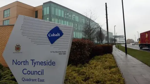 A sign for North Tyneside Council. It is grey with blue text. Behind the sign is the council building, one wall of which is full of glass panels and another is an orange brick. There are some bushes just behind the sign and a path to the right of it.