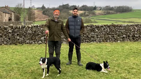 BBC Two men stand in a field, with two dogs in front of them. Behind them is a dry stone wall, and beyond that a farm house, rolling hills and power lines.
