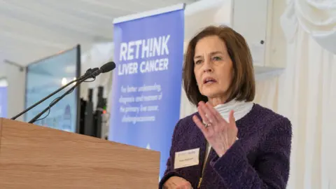 AMMF AMMF chief executive, Helen Morement gives a speech in front of a a purple banner that says "rethink liver cancer." She has brown bobbed hair and wears a white roll neck jumper under a purple zip-up jacket.