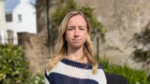 A girl with shoulder-length blonde hair, wearing a jumper with blue and white horizontal stripes. She is staring directly at the camera.