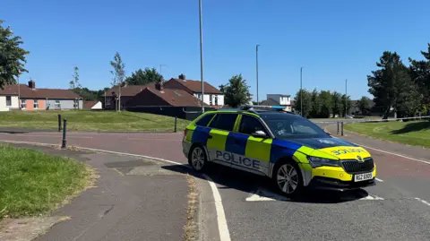 Pacemaker A marked blue and yellow police car is parked diagonally across a junction. There is police cordon tape just in the distance blocking off a road. There are some terrace houses to the left of the picture.