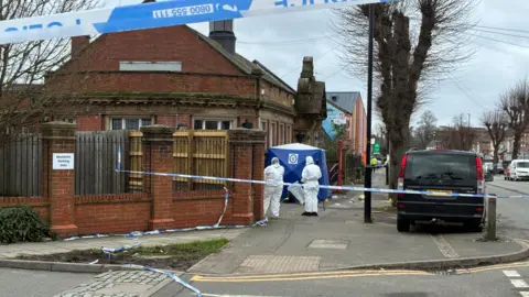 Side street view of Foleshill Library, a single-storey building dating from the Victorian era. There’s blue and white police tape restricting access to the street and a blue police tent erected at the library’s entrance. Two people covered in white head-to-toe PPE coverage stand aside the tent.