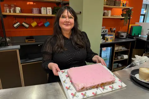 Sarah Farrell-Forster smiling into the camera as she holds a cake decorated with pink icing. She has long black hair and is wearing a black dress.